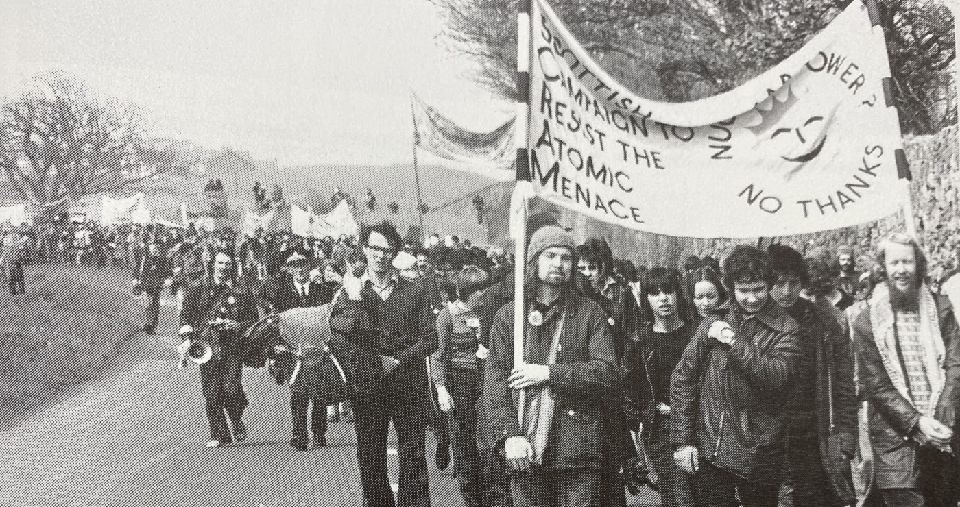 A black and white photo of a demonstration with people carrying a banner that says 'Scottish Campaign to Resist the Atomic Menace' and 'Nuclear Power? No Thanks' with a sun with a smiley face in middle. Source: 'Torness: Nuclear Power Station: From Folly to Fiasco' (Edinburgh: SCRAM, 1983) 