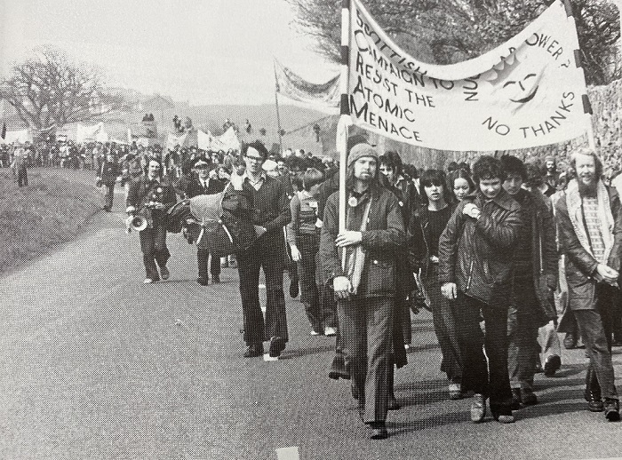 A black and white photo of a demonstration with people carrying a banner that says 'Scottish Campaign to Resist the Atomic Menace' and 'Nuclear Power? No Thanks' with a sun with a smiley face in middle. Source: 'Torness: Nuclear Power Station: From Folly to Fiasco (Edinburgh: SCRAM, 1983)'