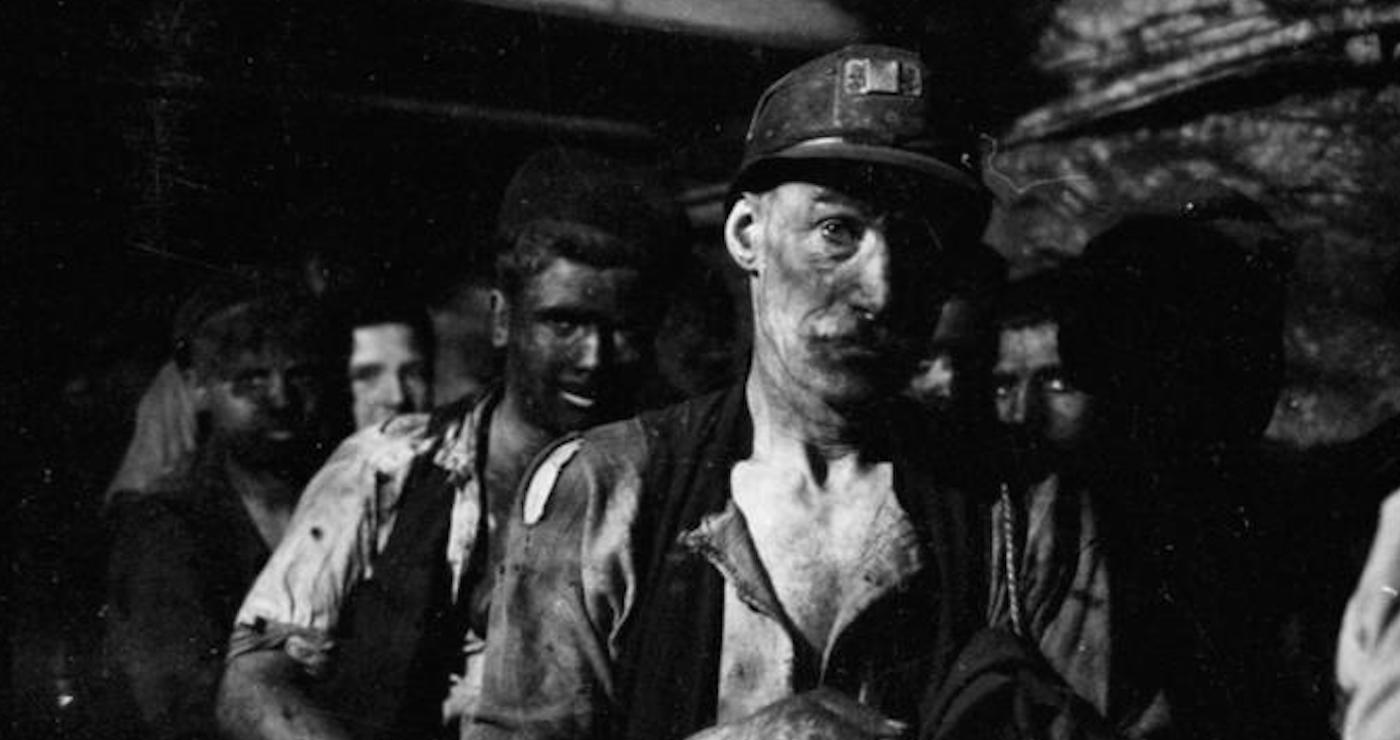 A black and white photo group of miners underground, walking forward led by a serious older man with a moustache wearing a helmet and holding a lamp. The face of the man behind is completely black with coal dust.Source: Ministry of Information, 1942, Imperial War Museum https://www.iwm.org.uk/collections/item/object/205199325