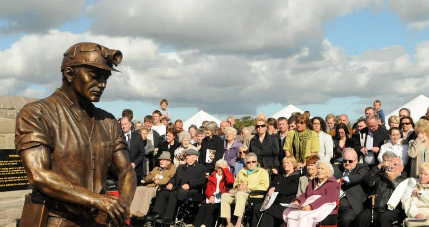 A statue of a miner leaning on a pickaxe with a crowd of people including older people in wheelchairs around it. Source / Copyright: https://www.facebook.com/Auchengeich-Miners-Memorial-916368968418660/photos	