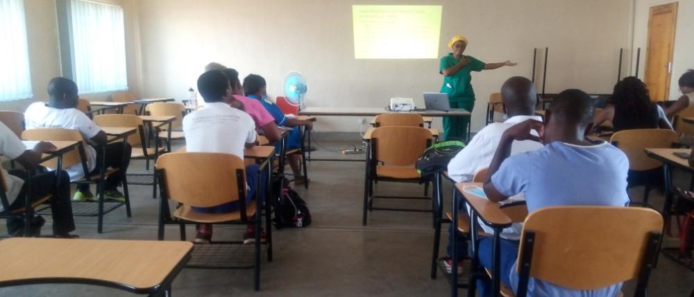 A woman in green scrubs and a yellow head wrap gestures to an audience in a classroom, with a powerpoint presentation behind her on the wall.