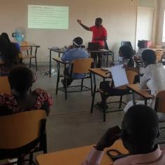 A man in a red shirt gestures to a powerpoint presentation in front of a classroom of observers