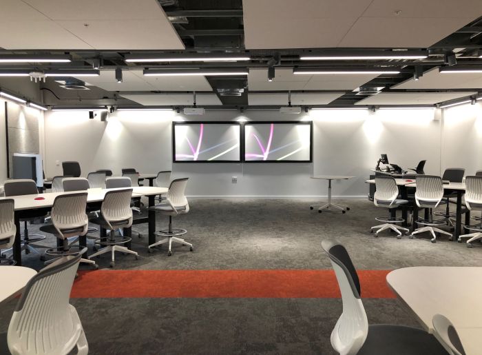 Flat floored teaching room with tables and chairs, PC, lectern, and two large screens. 