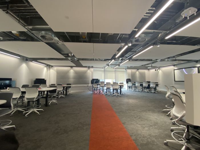 Flat floored teaching room with tables and chairs, and large screen.