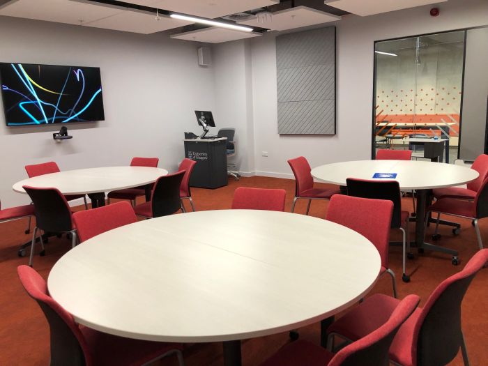 Flat floored teaching room with tables and chairs, PC, lectern, and large screen.
