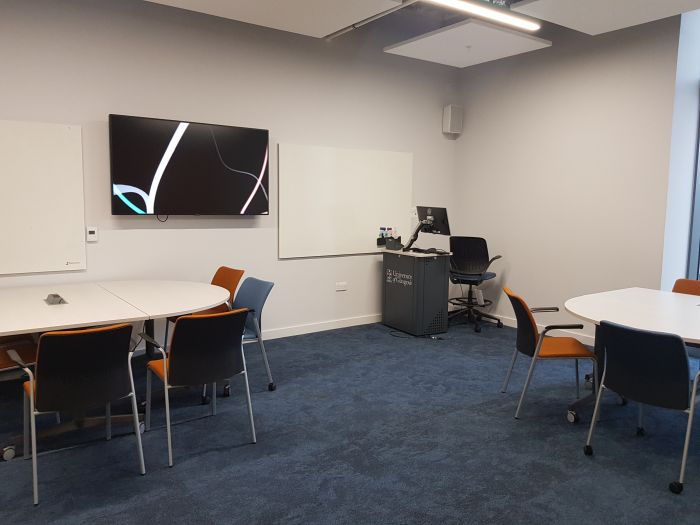 Flat floored teaching room with tables and chairs, PC, lectern, and large screen.