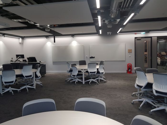 Flat floored teaching room with tables and chairs, PC, lectern, and two whiteboards..