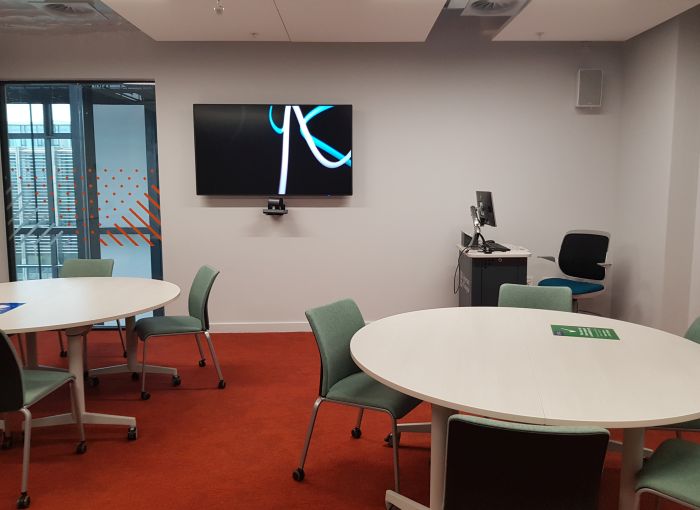 Flat floored teaching room with tables and chairs, PC, lectern, and large screen.