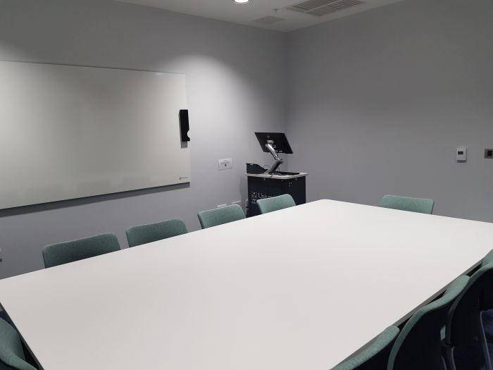 Flat floored teaching room with table and chairs, PC, lectern, and white board..