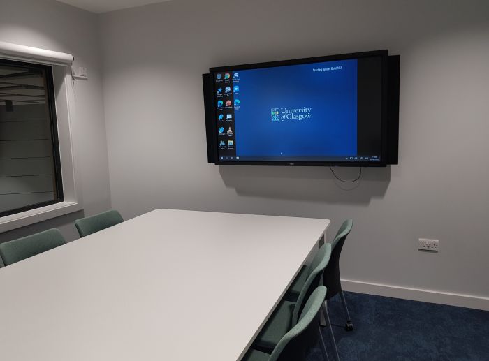 Flat floored teaching room with table and chairs, and large screen.