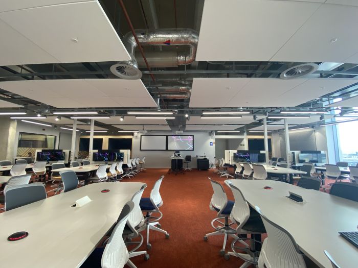 Flat floored teaching room with tables and chairs, PC, lectern, and two large screens.