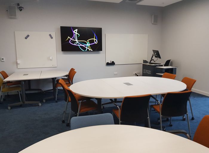 Flat floored teaching room with tables and chairs, PC, lectern, large screen and two whiteboards.