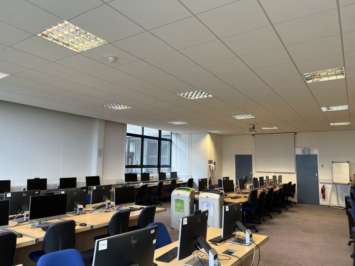 Flat floored teaching room with rows of PC and monitors, moveable whiteboard, large screen, and projector.