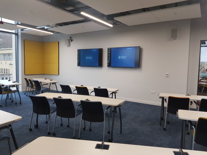 Flat floored teaching room with rows of tables and chairs, and two large monitors.