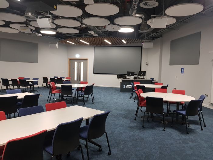 Flat floored teaching room with tables and chairs in groups, large screen, PC, and lectern.