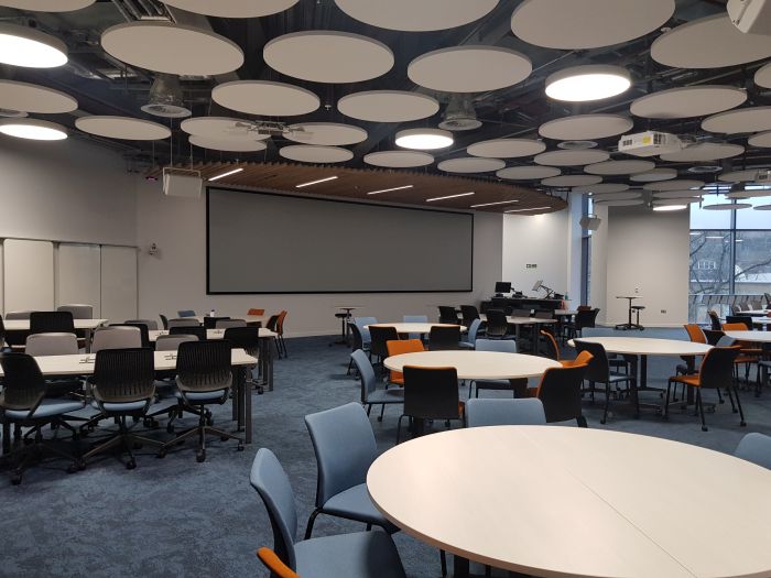 Flat floored teaching room with tables and chairs in groups, PC, lectern, large screen, and whiteboards..