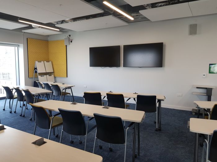 Flat floored teaching room with rows of tables and chairs, two large screens, and handheld whiteboards.