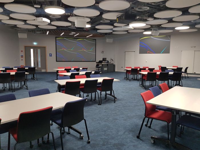 Flat floored teaching room with tables and chairs in groups, PC, lectern, two large screens, and whiteboards.