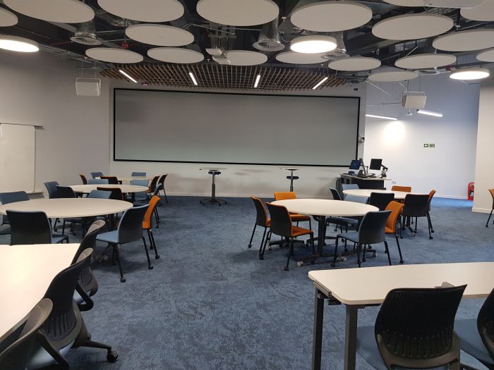 Flat floored teaching room with tables and chairs in groups, PC, lectern, large screen, and whiteboard.