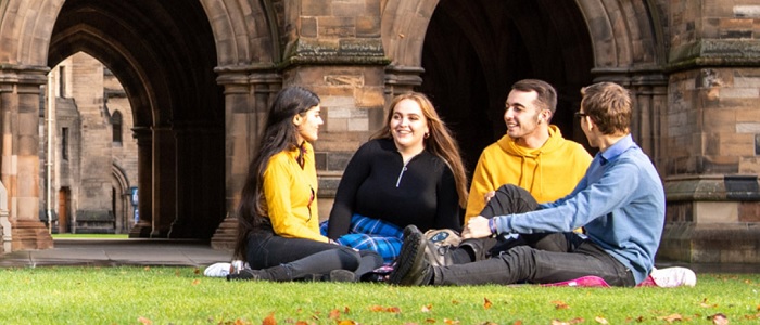 Group of four students sitting on the grass in Glasgow University quadrangle with the cloisters behind them. Source: GU Image Library