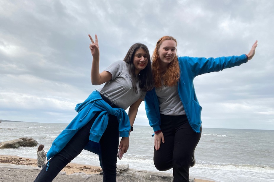 Two Residence Life Assistants wearing grey t-shirts and blue hoodies posing for a photo at the beach