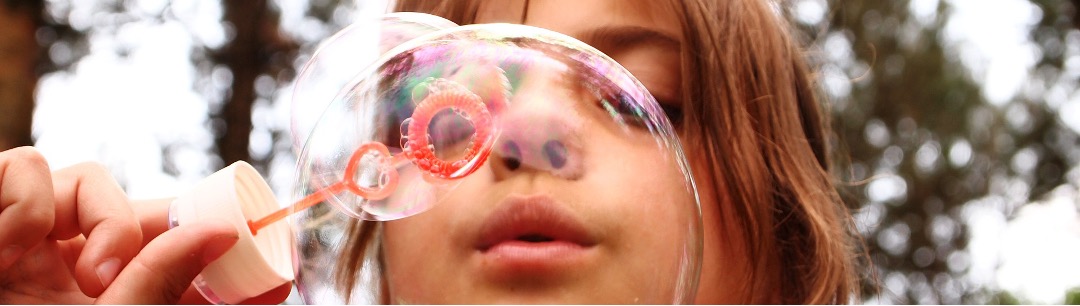 A young child blows a bubble towards the camera with a bubble wand. 