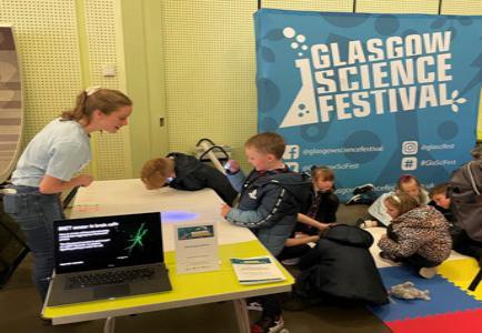 Photograph of a GSF in Action student and 2 children using UV lights to reveal invisible ink. On the mat on the floor more children are drawing and using the UV light. 