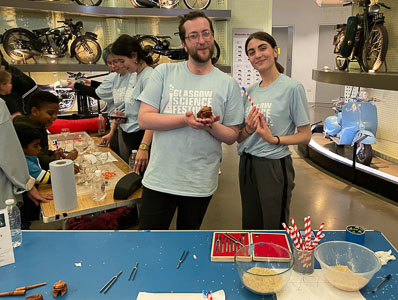 Photograph of two GSF in action students holding a musical frog and a homemade 'rattlesnake' whilst in the background two more students are demonstrating sounds in water to two children with wine glasses filled with different amounts of water. 