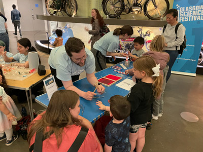 Photograph of a GSF in Action student demonstrating using different tools to make sounds to an adult and two children, in the background another student is helping three children make rattlesnakes out of paper straws. 