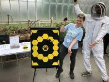 Photograph showing two GSF in Action students with a bee poster board mad of hexagons to simulate a beehive. One of the students is in a beekeepers suit. 