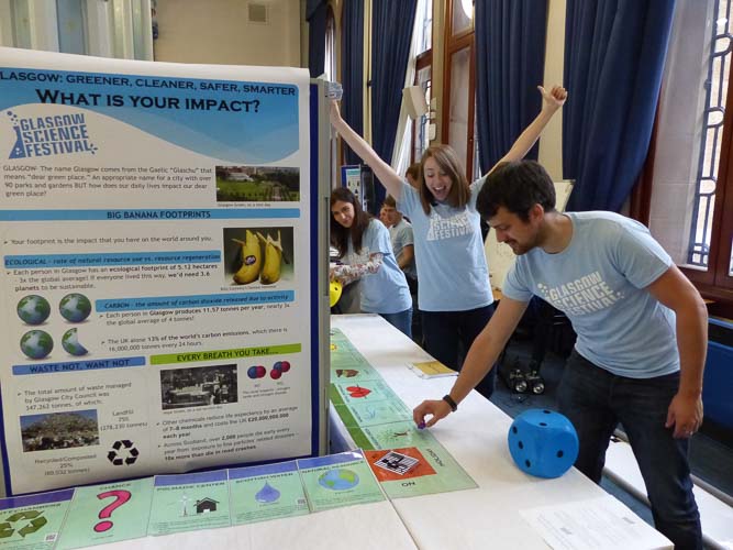 Photograph showing three GSF in Action students with a large monopoly style game board. There is a large poster discussing individual carbon footprints. 