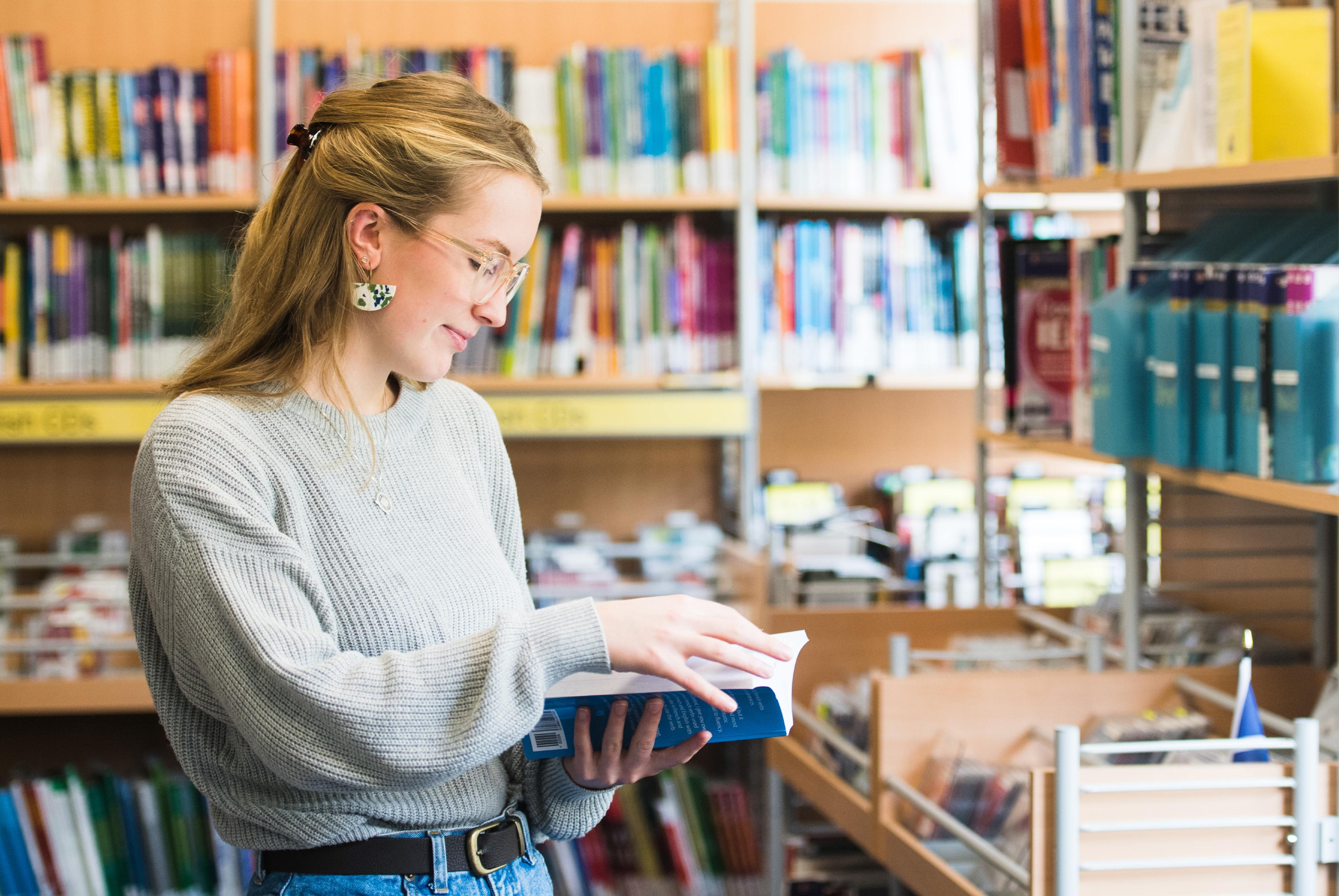 A side on view of a student looking at a journal in a library setting. with books on shelves in the background