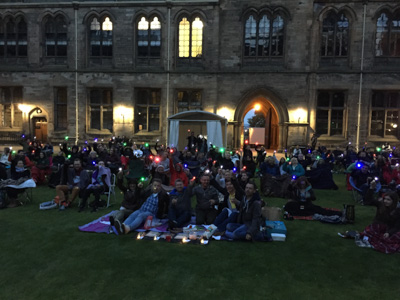 Photograph showing a large group of people sat outside the University of Glasgow holding up lights and ET fingers whilst watching a film. 