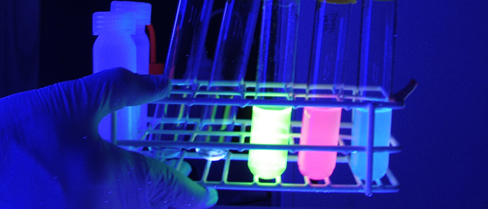 Researcher's gloved hands holding a tray of luminescent test tube vials in a darkened room