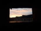 A photo shot through a small porthole-like window looking out onto a sunset sky over a low and darkened landscape.