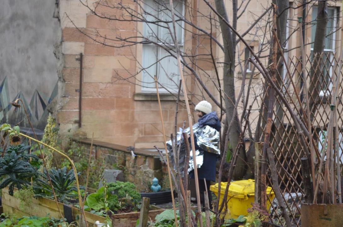A distant photo of one of the workshop participants wearing one of the reflective blankets in a community garden reflecting on their experience during the winter. 