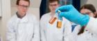 A hand in a blue latex glove holding two small vials with coloured liquid inside. In the background are people wearing white lab coats  Source: Glasgow University image library Dumfries lab 06