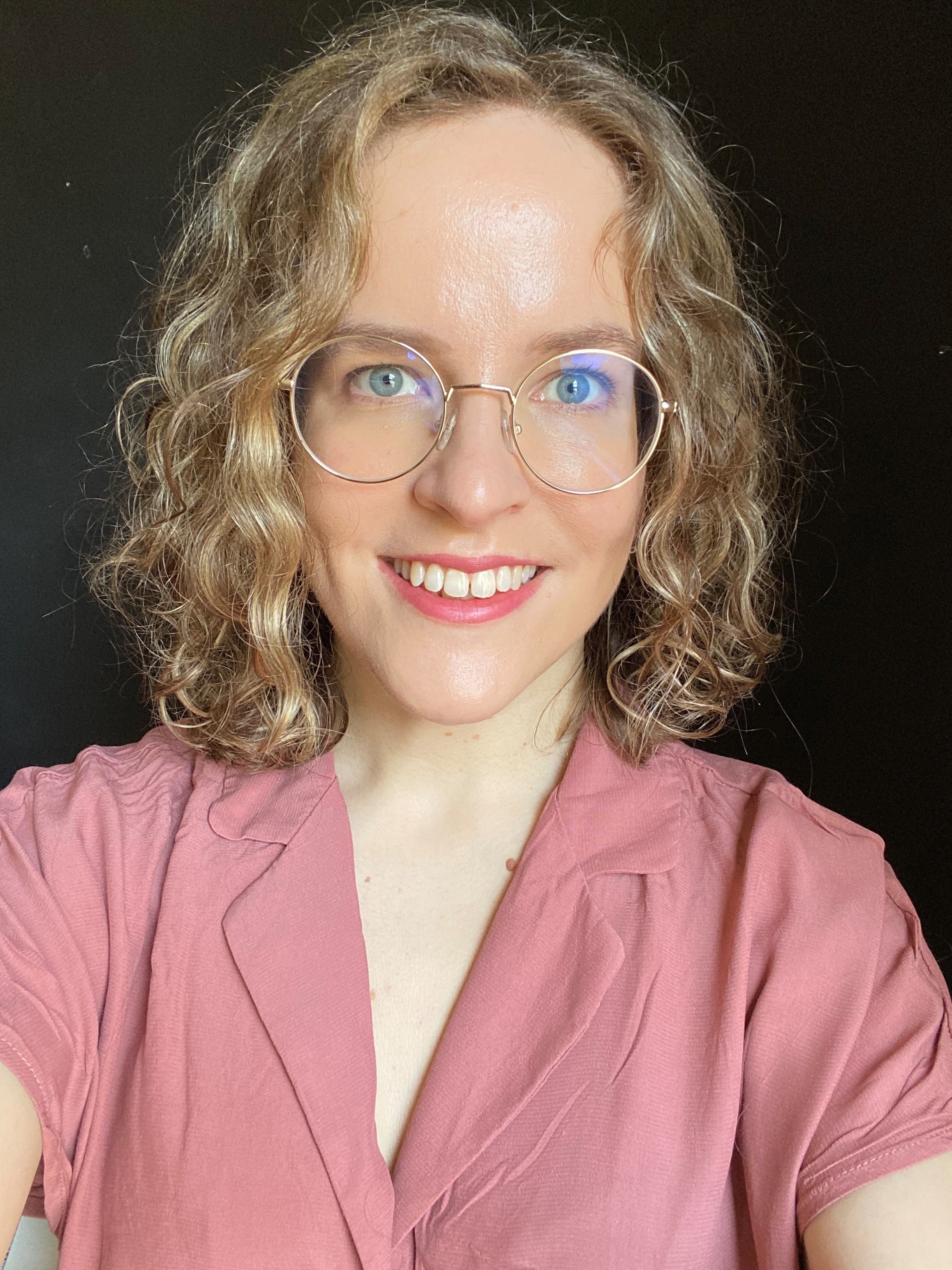 Picture of Ida who has curly hair and round glasses, smiling into a camera with a black background wearing a pink shirt