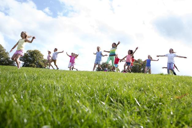 A group of happy looking children run along the grass with their arms outstretched. The sky is blue and there are some trees in the background.