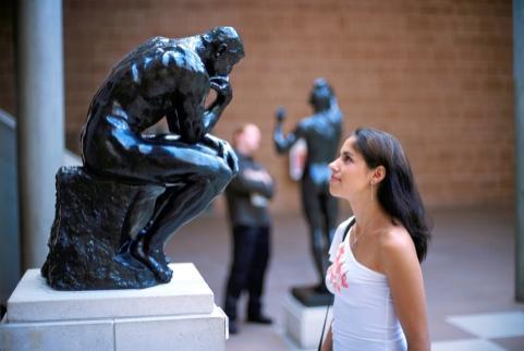 photograph of a young woman with long black hair and a white top off the shoulder looking up at a black statue of a man hunched with his hand to his chin, on top of a white pedestal. In the back, another person looking at another black sculpture, against a brick-wall background