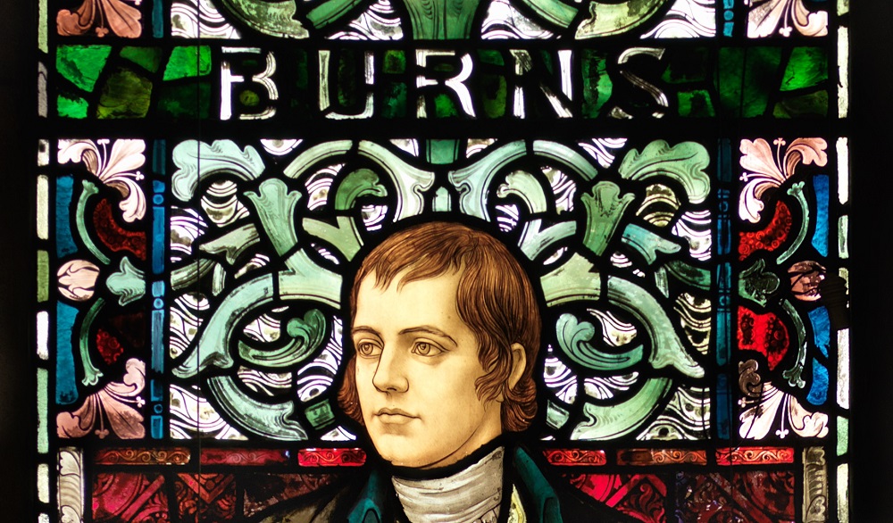 Stained glass window with a portrait of Robert Burns on a floral background and the name 'Burns' in the window above his head. Source: University of Glasgow https://www.gla.ac.uk/events/globalburns/ 
