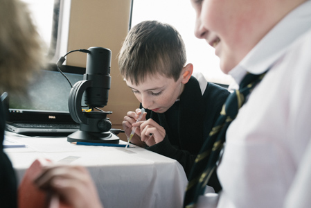 Photograph of a boy holding a pipette and dropping some water sample onto an empty glass microscope slide. He is sitting in front of a microscope. 