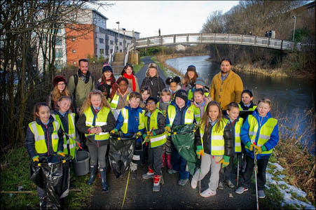 Photograph showing a group of school children with their teachers. They are all wearing hi-vis vests and holding litter pickers or bin bags. 
