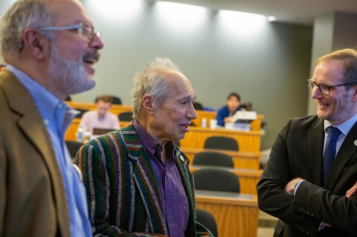 Sam Peltzman with Graeme Roy and another person beside them chatting in the centre of a lecture hall. Close-up on Sam Peltzman's animated face and raised hands while he is speaking to a person whose face can't be seen. Source: Chicago Booth University