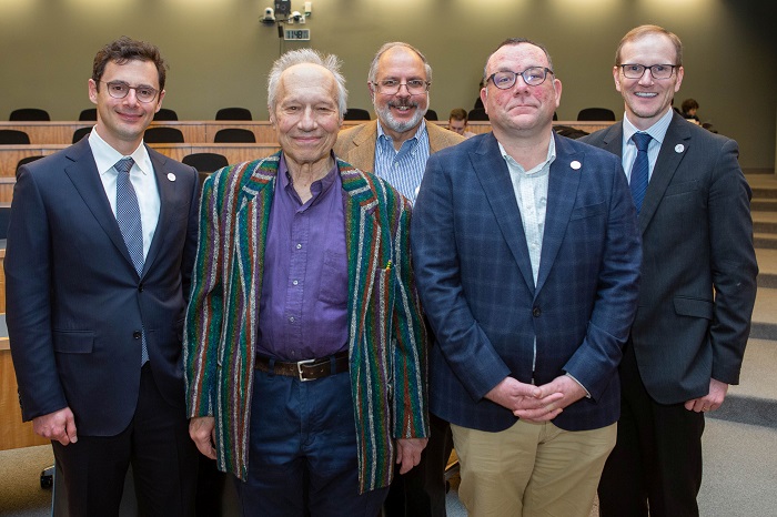 A group photo with Sam Feltzman, Tom Scotto in front and Graeme Roy and two more men standing behind them Close-up on Sam Peltzman's animated face and raised hands while he is speaking to a person whose face can't be seen within a lecture hall. Source: Chicago Booth University