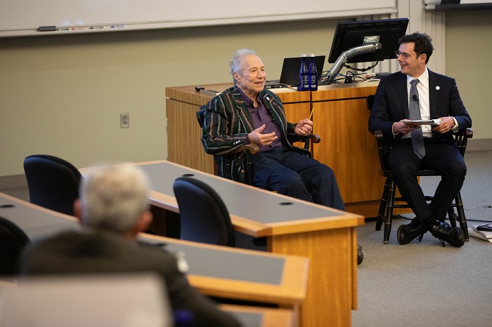 Sam Feltzman Sitting in a lecture theatre presenting to an audience around the theatre with another man next to him. We see behind the audiences in front Close-up on Sam Peltzman's animated face and raised hands while he is speaking to a person whose face can't be seen. Source: Chicago Booth University