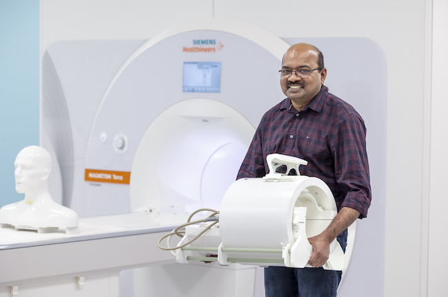 Dr Shajan Gunamony holds the 7T Head Coil beside the 7T MRI scanner
