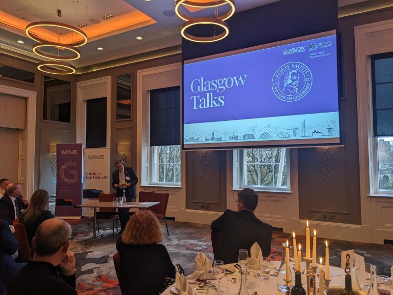 An event with groups of people at round dinner tables with white tablecloths and candles listening to Glasgow University Principal Anton Muscatelli. A screen with the text 'Glasgow Talks' and 'Adam Smith 300 Anniversary 1723, 2023' is at the front of the room. Source: University of Glasgow