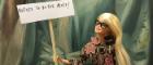 A blonde barbie doll wearing black glasses and a black patterned coat with gold braiding and pink embroidered flowers holds up a mini placard that reads 'Refuse to be the Muse' in front of a green painted background, which appears to be a section of a large painting 