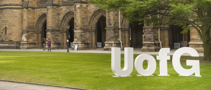 Large white letters spelling out UofG on the grass of gilbert scott quadrangle with the cloisters in the background